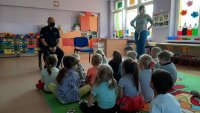 Policjant prowadzi zajęcia z grupą dzieci w przedszkolu. Grupa dzieci widoczna jest tyłem, siedzą na przeciwko policjanta. Obok stoi opiekunka.