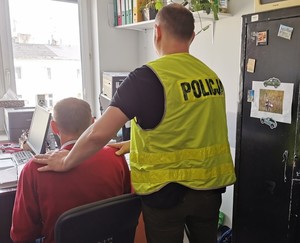 mężczyzna siedzi na krześle w pokoju za nim stoi policjant