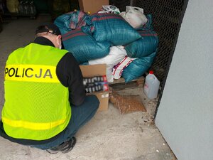 Policjant w kamizelce liczy zabezpieczone papierosy. W tle widoczne worki z zabezpieczonym tytoniem.