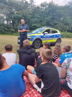 policjant rozmawia z dziećmi podczas spotkania profilaktycznego stojąc w lesie przy radiowozie.