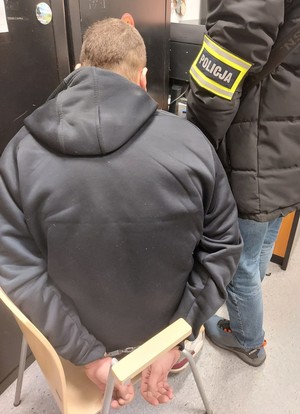 Zatrzymany mężczyzna siedzi w pokoju a obok niego stoi policjant.